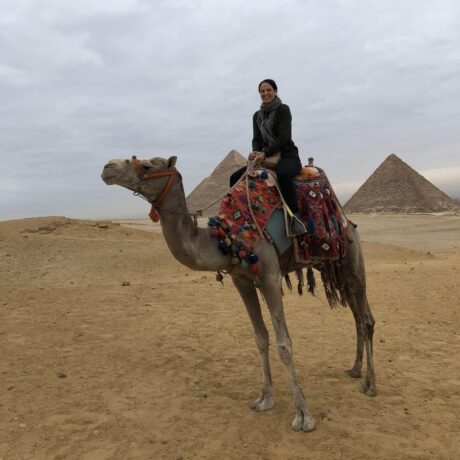 Kirsty Nielsen - Founder of Zen Aroma smiling while seated on a camel in front of the great pyramids of Giza, with a cloudy sky backdrop. The camel is decorated with colorful traditional saddle blankets.