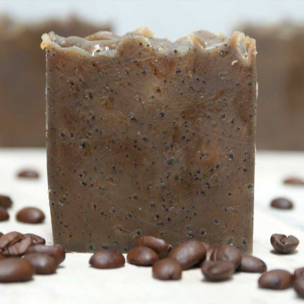 A close-up of a homemade coffee-infused soap bar, textured and dark brown, standing upright on a white surface surrounded by scattered coffee beans.